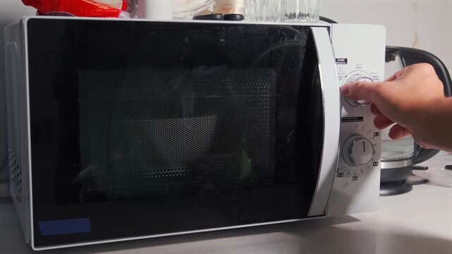 Hand turning rotary control on microwave oven, selecting heating mode and time, kitchen appliance operation, close-up of dial adjustment during food preparation at home