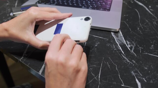 Hand removing SIM card from smartphone slot using eject tool, close-up of mobile device maintenance, user accessing SIM tray for replacement or repair on desk near laptop