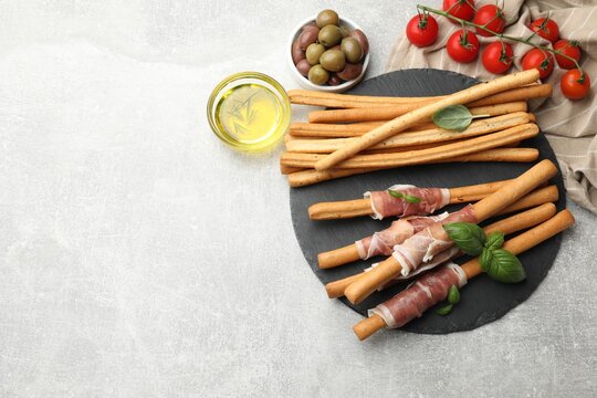 Grissini breadsticks with prosciutto, tomatoes, oil, olives and basil on light table, flat lay. Space for text