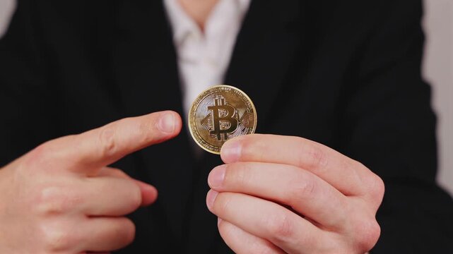 Une main montre une pi&egrave;ce de Bitcoin. En arri&egrave;re plan, une personne en costume et chemise blanche.