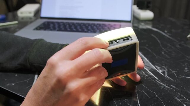 Hand rotating control knob on portable RGB lamp adjusting brightness or color settings, close-up of lighting device control, user changing parameters indoors on desk near laptop