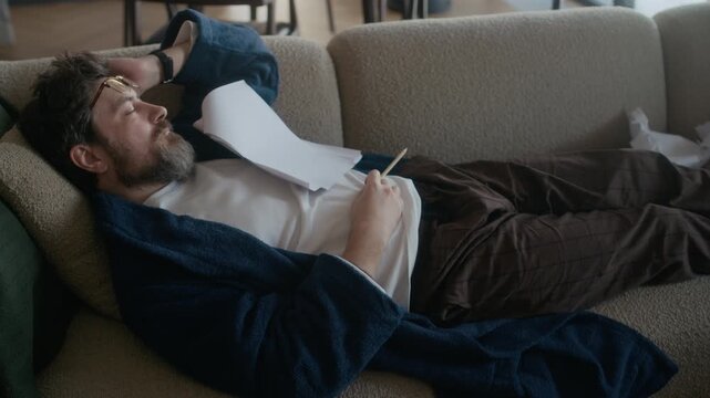 Mature adult Caucasian male writer wearing robe sleeping on sofa with novel draft on his chest, holding pencil, recharging after long work period