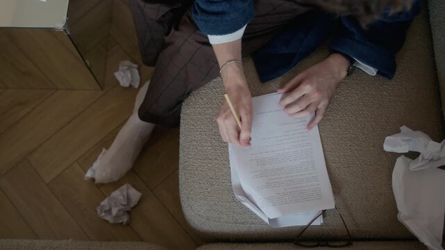 From above view of unrecognizable man sitting on sofa writing notes in novel draft paper, crumpled papers scattered nearby