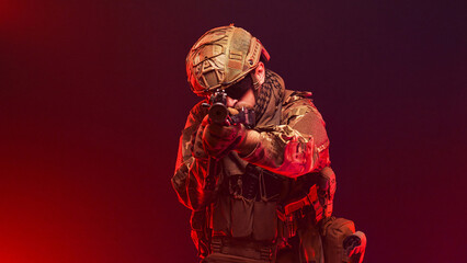Naklejka premium Military fighter in tactical gear with rifle on red smoky studio background. Concept combat and defense. 