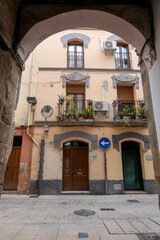Naklejka premium Historic residential building in Plasencia, Spain, featuring traditional balconies with plants and ornate stone lintels, framed by a medieval archway in the heart of the old city center.