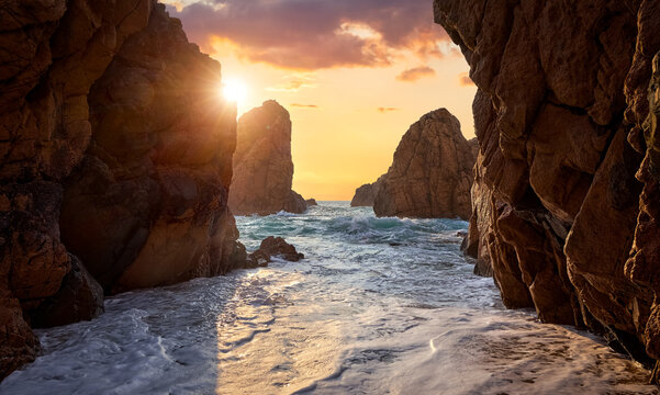 Sand beach among rocks on evening sunset. Ursa Beach near Cape Roca (Cabo da Roca) at Atlantic Ocean coast in Portugal. Summer landscape. Beach with splashes of waves crashing of stone rocks.