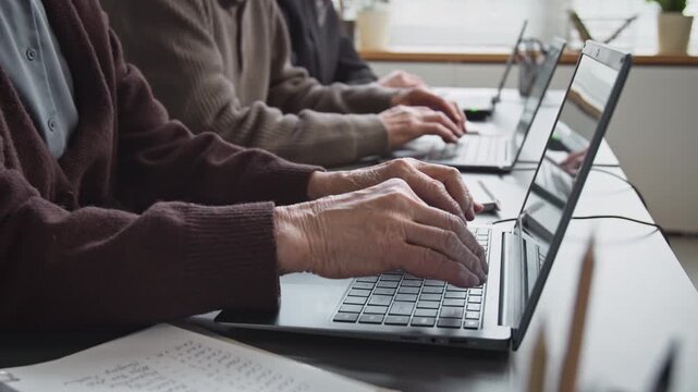 Side view close up shot of wrinkled hands typing on keyboard while group of senior people using laptops sitting in row at desk and doing tasks during computer literacy class, copy space