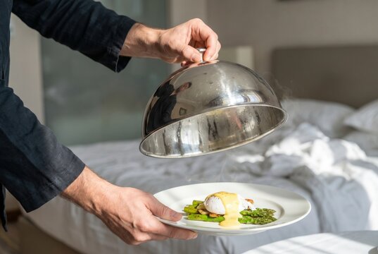Waiter hand lifting a silver cloche to reveal a hot breakfast plate of eggs benedict in a hotel room