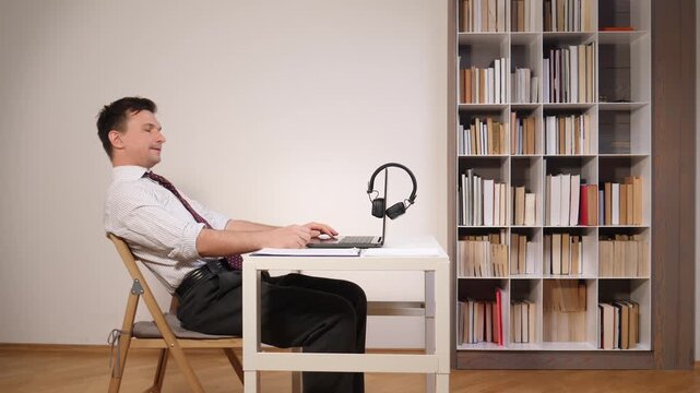 Male professional working remotely from home or office, typing on a laptop with hands stretched forward, displaying poor ergonomic posture at a desk with headphones and a bookshelf in the background
