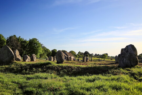 Antike Menhire erstrecken sich in langen Reihen &uuml;ber eine gr&uuml;ne Wiese vor einem einsamen Steinhaus unter strahlend blauem Himmel im warmen Abendlicht der Bretagne.