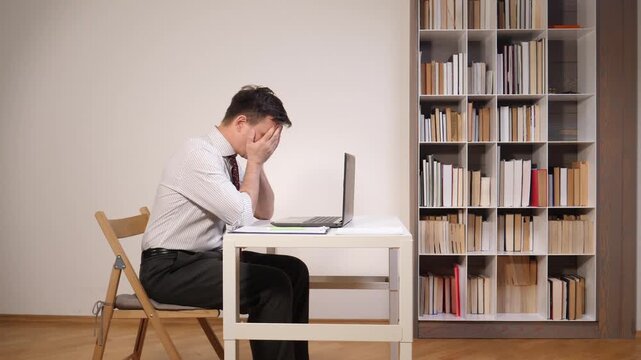 Businessman covering his face with hands while sitting at a desk with a laptop, feeling overwhelmed and frustrated by work and experiencing professional burnout