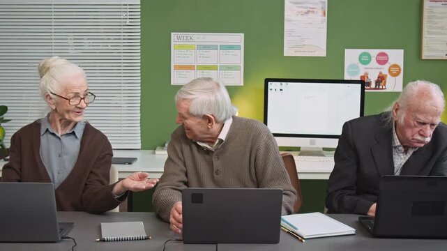 Front view shot of group of senior people using laptops while sitting in row at desk in computer literacy class and chatting cheerfully discussing tasks