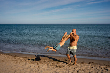 Fototapeta premium Father swinging son by the arms on sandy beach, joyful family bonding moment by the sea, shirtless man playing with child in swim shorts during summer vacation at seaside