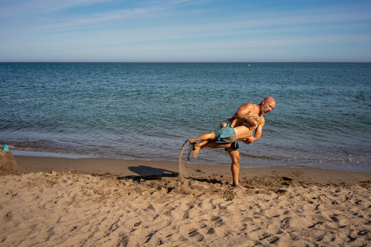 Father lifting and spinning his young son playfully on sandy beach by the sea, both covered in sand, enjoying active fun, bonding, and joyful summer vacation under bright blue sky