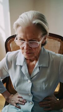 Mature woman in light blue shirt holding stomach in pain sitting on cane chair indoors medium shot