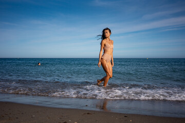Fototapeta premium Fit young woman in beige bikini walking along seashore with wet skin glowing in sunlight, looking away confidently on summer beach with blue ocean waves in background