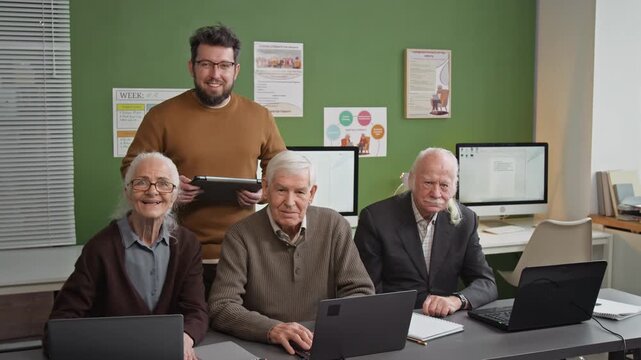 Front view shot of group of senior people using laptops while sitting in row at desk in computer literacy class posing with friendly male teacher, all looking at camera smiling