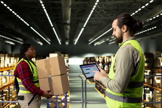 Male employee checking parcel status on a tracking software with tablet, shipment details and AWB tags ensuring package tracking in warehouse. Adult man wearing safety vest on platform.