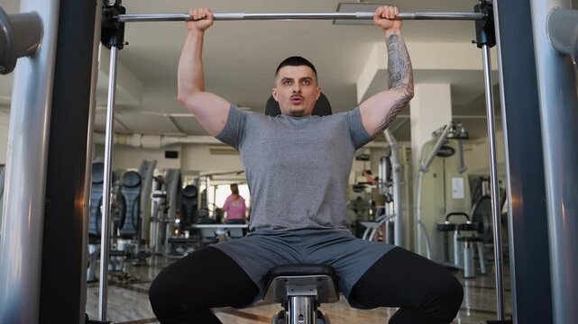Muscular tattooed athlete doing shoulder press on smith machine