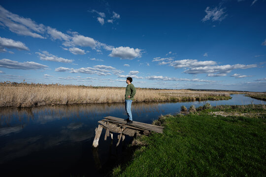 Man standing on an old wooden dock by a calm river looking across reed marshes under a wide blue sky in a peaceful rural landscape