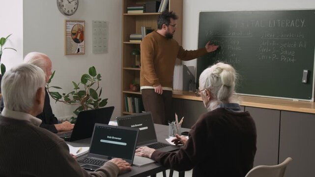 Scene of male teacher giving computer literacy class to group of senior people sitting at table together with open laptops and listening intently, community outreach program