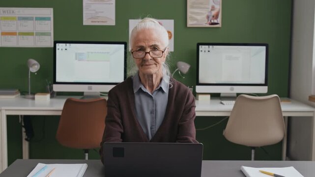 Front view shot of white haired senior woman using laptop and looking up at camera with smile while sitting at desk alone in library or computer class, copy space