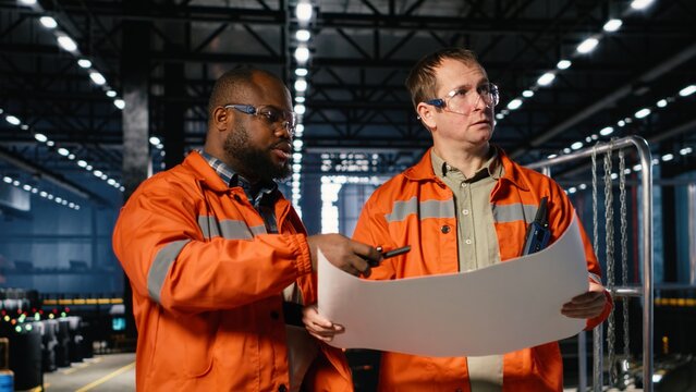 Two supervisors use plans to oversee construction and production tasks, surrounded by industrial machinery, metal tools and engineering elements that support plant operation.