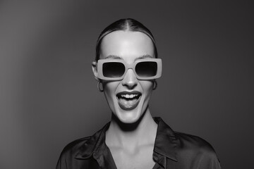 Fototapeta premium White woman with light skin wearing large sunglasses laughs with open mouth on a grey studio background. Concept of fashion, joy, and confident mood.