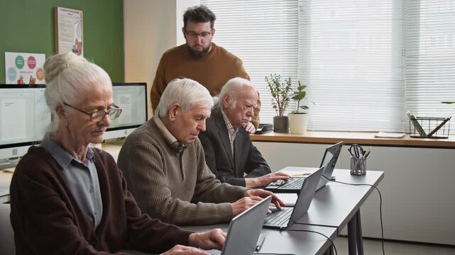 Side view shot of group of senior people using laptops and doing tasks online while sitting in row during computer literacy class, with male teacher assisting overseeing work, copy space