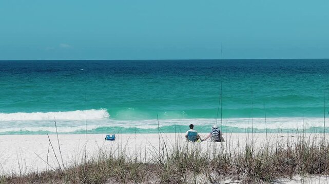 wide shot of couple sitting on white sand beach facing turquoise ocean