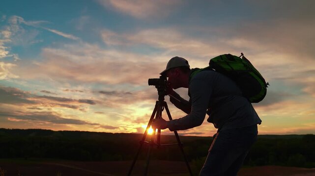 Traveler taking pictures beautiful natural landscapes. Male photographer with backpack uses camera tripod, nature, sky sunset. Traveler with tripod camera takes beautiful landscape shots. Silhouette.