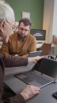 Vertical shot of friendly bearded man teaching computer literacy to senior woman and showing how to turn on laptop pointing at power button