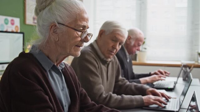 Tilt up shot of white haired senior woman using laptop in computer literacy class and wearing glasses while sitting in row at desk with group of seniors , copy space