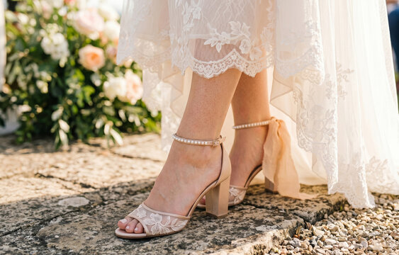 Low angle view of a bride's feet wearing beautiful lace block heel sandals with pearl straps on a stone path.