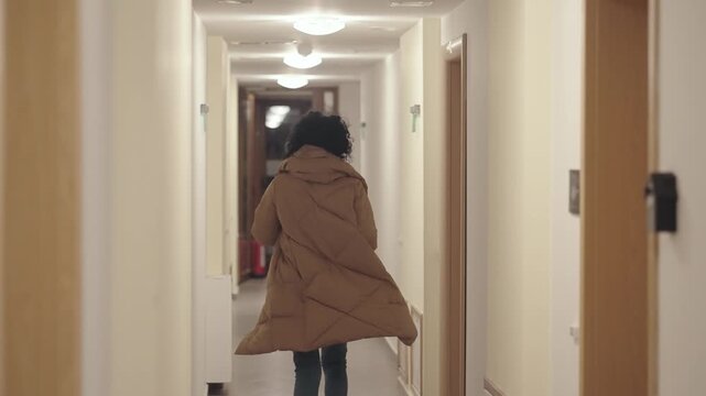 Curly hair hispanic woman running in building corridor dressed in winter jacket ang blue jeans, slow motion