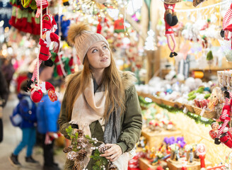 Fototapeta premium Young woman walks view hanging toys near window of New Year souvenirs seller, choose ornaments for Christmas tree. New Years rush. Shes fiddling with garland of fir cones..