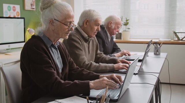 Side view shot of group of senior people sitting in row and using laptops typing on keyboards and doing tasks during computer literacy class, focus on white haired woman in foreground, copy space