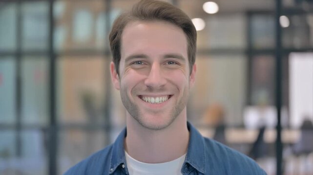 Close-up portrait of a smiling young man with short brown hair, looking directly at the camera in a professional office setting