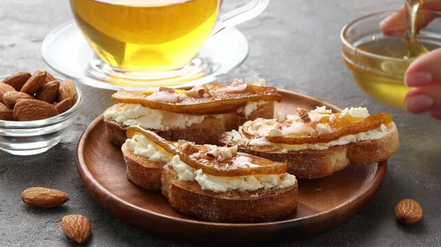 Woman dripping honey onto delicious bruschettas with cream cheese, pear and almonds at grey textured table, closeup