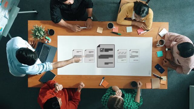 A top-down view of a diverse team engaged in a business meeting. Individuals discuss plans and analyze documents on a wooden table in a modern office environment. Raster