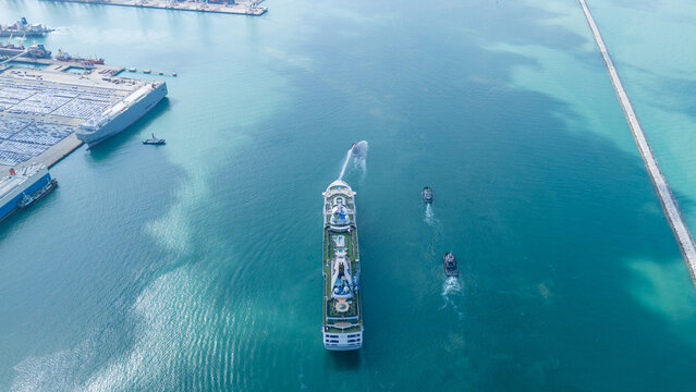 luxury cruise ship escorted by three tugboats in clear turquoise ocean water. One tugboat performing a ceremonial water salute. Travel, tourism, and maritime leisure.