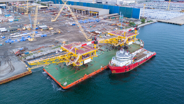 offshore oil and gas platform structure loaded on a heavy transport barge with a supply vessel at a coastal shipyard. Global energy industry and marine construction.