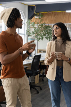 Diverse coworkers chatting while holding coffee cups, wearing beanie and blazer in open-plan office