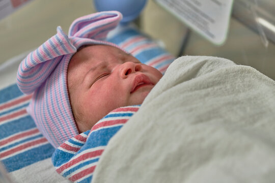 A newborn baby sleeping in a bassinet while swaddled and wearing a cap and bow.