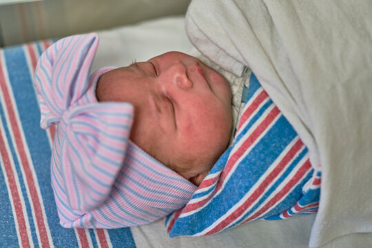 A newborn baby sleeping in a bassinet while swaddled and wearing a cap and bow.