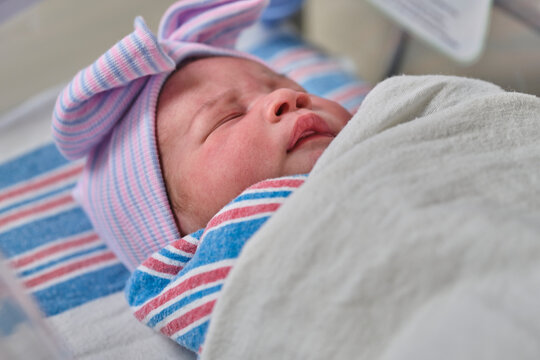 A newborn baby sleeping in a bassinet while swaddled and wearing a cap and bow.