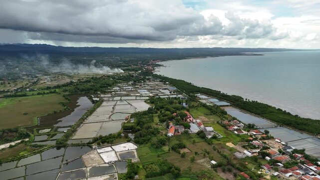 Aerial perspective of a serene coastal landscape, highlighting a vibrant fishing village and extensive salt pans under a dramatic sky