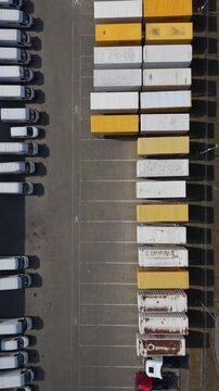 Vertical screen Aerial View Container Trailer Yard Rows Sunlit Industrial Depot With Parallel Lines Of White And Yellow Containers, Parked Trucks Flanking Central Aisle, Gritty Asphalt