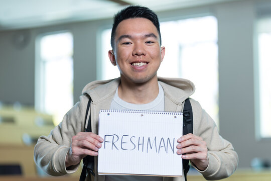An Asian young man smiles while holding a Freshman sign in a university lecture hall, perfect for freshman orientation, university enrollment, or first-year student concepts.
