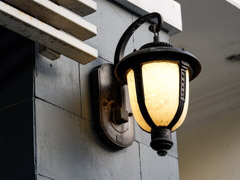 A classic black metal wall lantern with a warm glowing glass shade mounted on a gray stone wall. This decorative outdoor lighting fixture provides elegant illumination for building exteriors.
